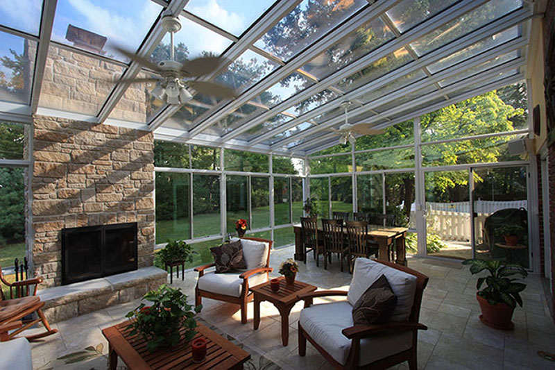 A large sunroom with a fireplace and ceiling fans furnished with comfortable wood furniture and plants.  