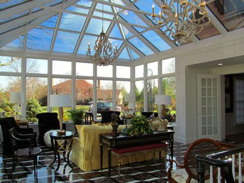 A conservatory interior with tile floors, chandeliers, sitting room furniture, and windows all along the roof and back wall.